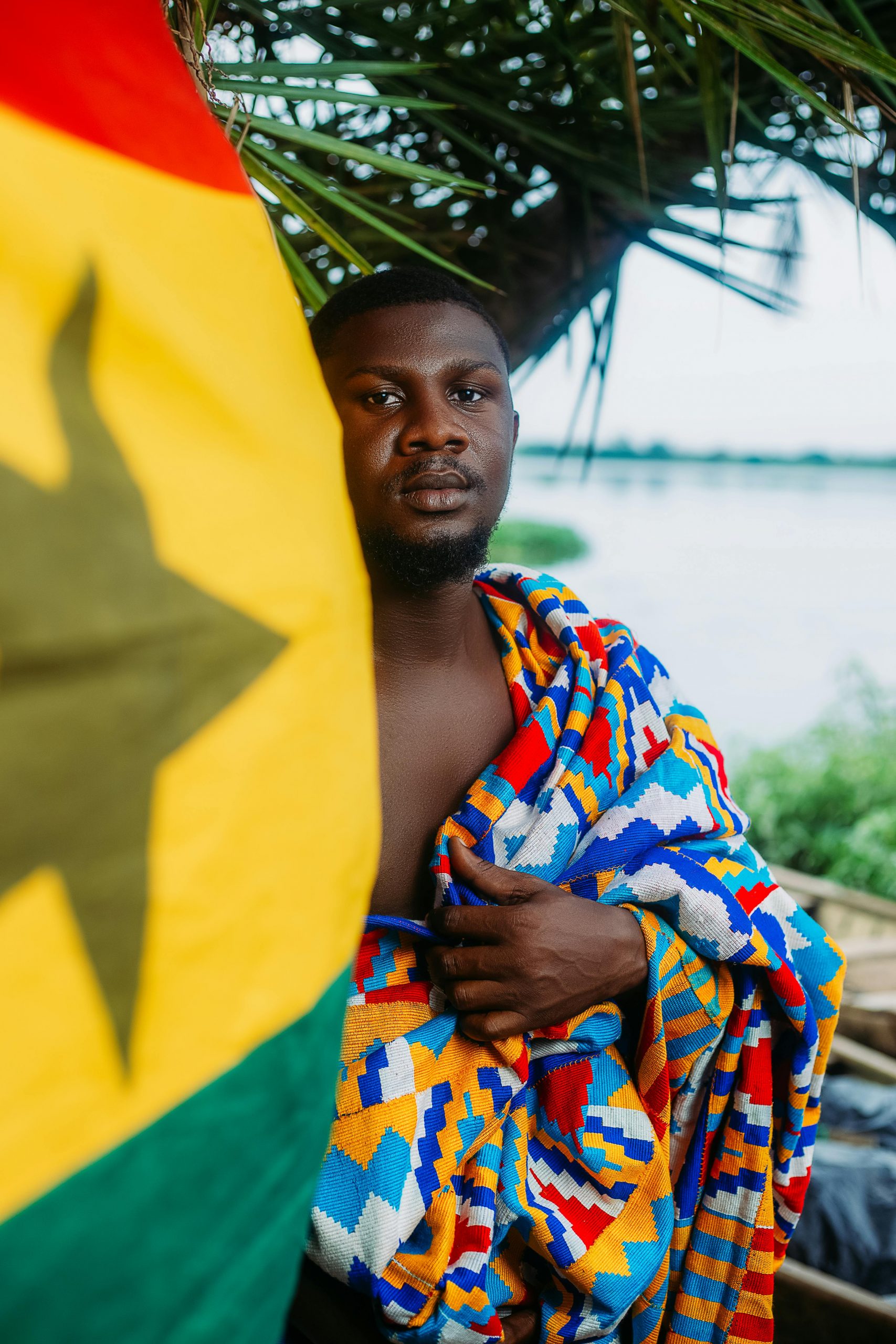Man in vibrant Kente cloth with Ghana flag by a riverside.